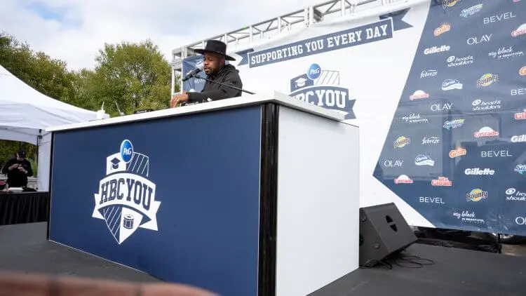 A black man wearing a black hat and sweater stands on stage at a turntable, as he plays music during a P&G HBCU homecoming season event.