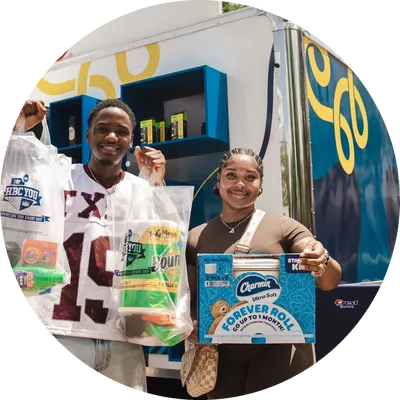 A black male and female stand outdoors in front of a truck. They each hold up bags and boxes of various personal care products.