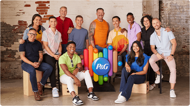 A group of twelve people are smiling for a group photo. In the middle of the picture is a rainbow-colored heart with the P&G logo in the center.