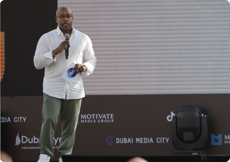 Man standing and talking into a microphone. He is wearing a white shirt and green trousers.