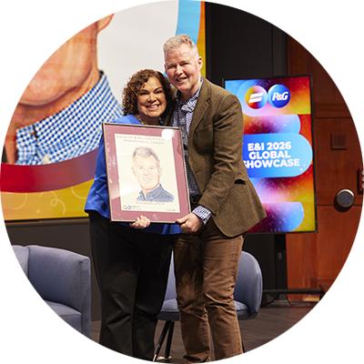 A woman with brown curly hair and blue jacket, and a man with short, light-colored hair in a brown jacket pose on a colorful stage. Between them, they hold a mounted and framed illustration of the man with the award year.