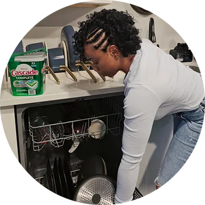 A woman with braided hair loads dishes into a dishwasher, with a tub of Cascade Complete detergent visible on the counter