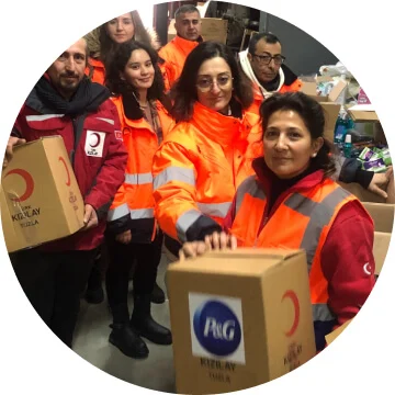 Seven people in orange safety jackets are holding boxes with the P&G logo on it. They are smiling towards the camera.