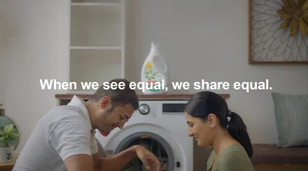A man and woman sit in front of a washer. White text in center says, "When we see equal, we share equal."