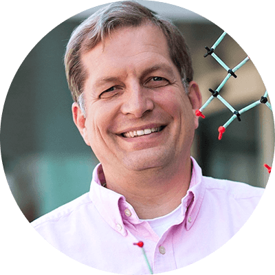 A man with short blonde hair wears a pink buttoned shirt. He smiles as he holds up a plastic molecular model.