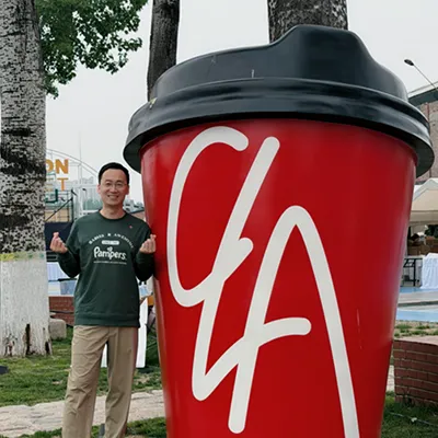 A man in glasses and a "Pampers" shirt stands smiling next to a huge red coffee cup. He gestures with his hands in front of trees and grass outdoors.