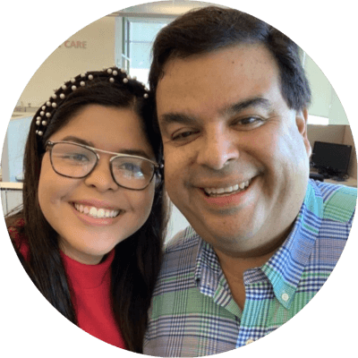 A Hispanic male with short dark hair and young Hispanic woman with long dark hair pose together as they smile. They stand in an office, with the words "home care" displayed on the wall in the background.