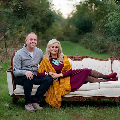 A smiling couple sits on a fancy white couch with carved wood details, placed in a green, natural outdoor setting with a path in the background.