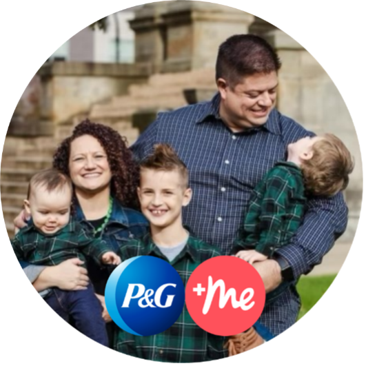 Woman with dark curly hair, stands holding her youngest son, while her husband on the right holds another son who is looking up at him. Their oldest boy stands between them. All are wearing blue plaid shirts and smiling.