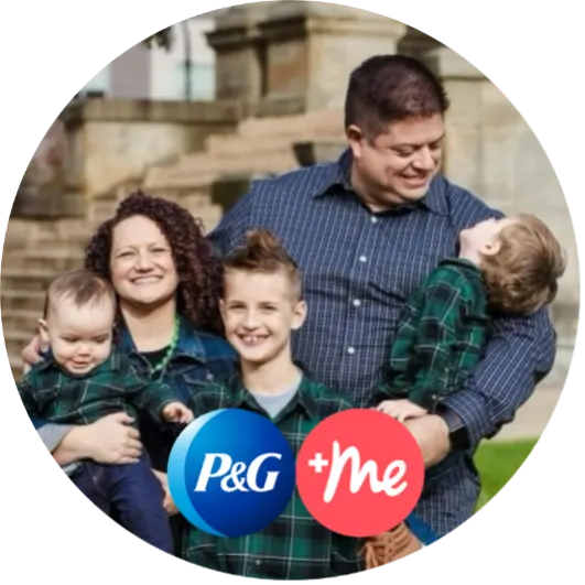 Woman with dark curly hair, stands holding her youngest son, while her husband on the right holds another son who is looking up at him. Their oldest boy stands between them. All are wearing blue plaid shirts and smiling.