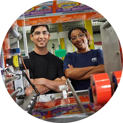 A male and female employee are in a manufacturing plant. They smile directly at the camera through some of the machinery.