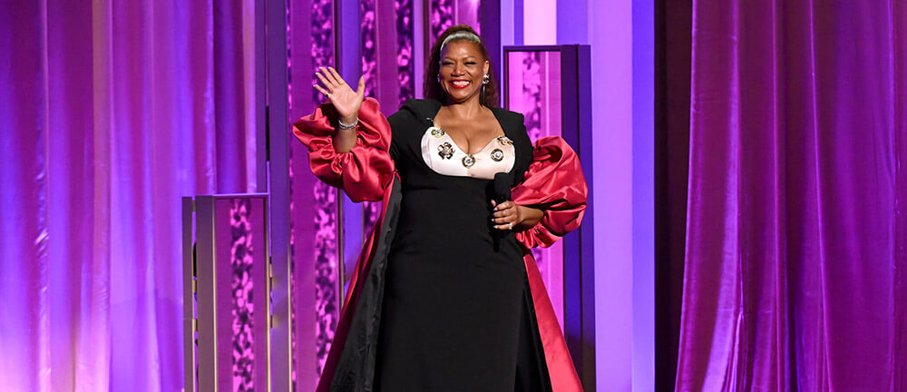 A Black woman in a formal black dress stands on stage with a microphone in her hand, smiling and waving at the camera.