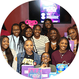 A group of seventeen young black college women gather and smile in front a table displaying Tampax period products and packaging.