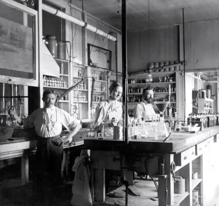 Two white men in white shirts and dark pants and one white woman in a white shirt and a dark skirt stand inside an industrial laboratory. In the black and white photo, they are surrounded by lab flasks and other materials.