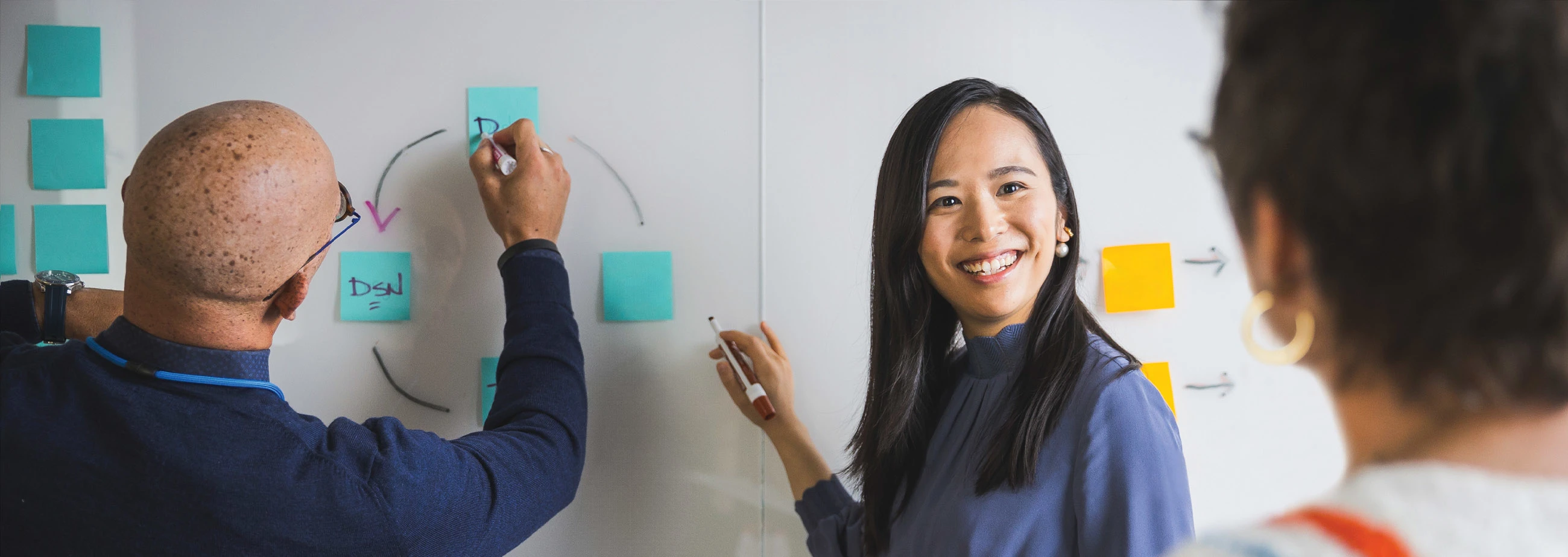 Man and woman writing on whiteboard while another woman looks on, her back to the camera.  