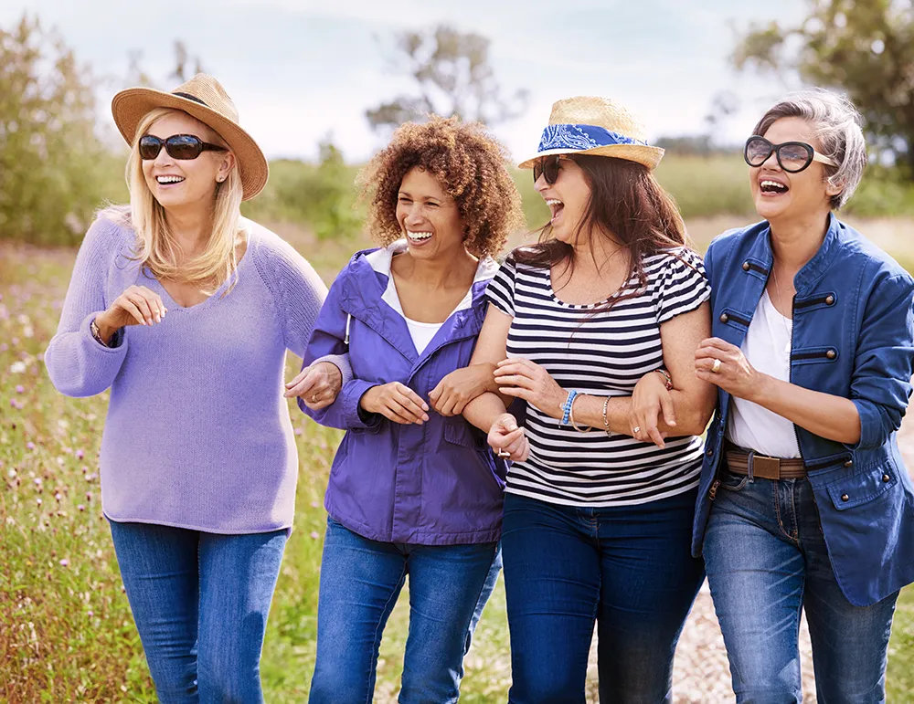 Four women walk arm in arm outdoors on a sunny day, laughing together. They are casually dressed in clothes of various shades of blue.