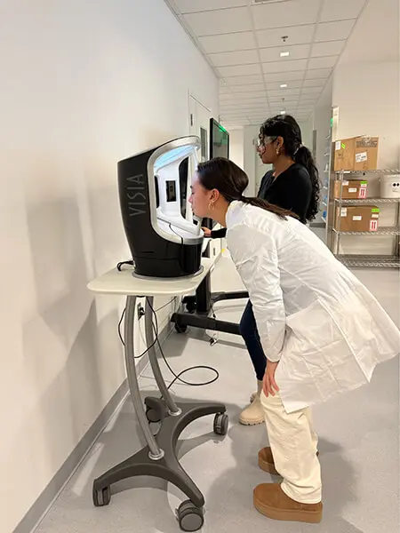 A young woman in a white lab coat leans into a machine to take her photo. In the background, a woman in lab goggles and a black shirt operates a computer screen.