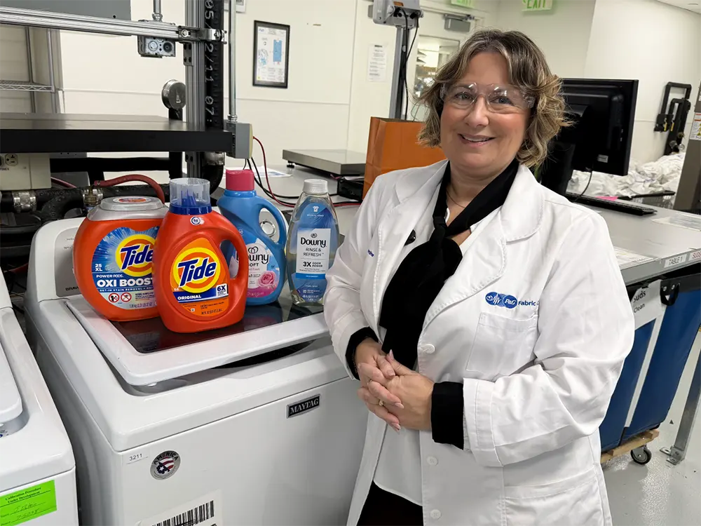A woman in a lab coat stands next to Tide detergent and Downy fabric enhancer placed on a washing machine in a lab.