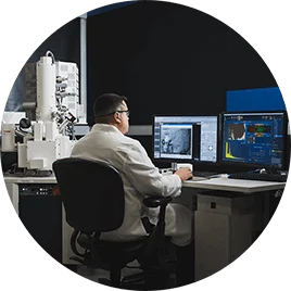 A man in a white lab coat sits at a desk in his lab, examining multiple computer screens.