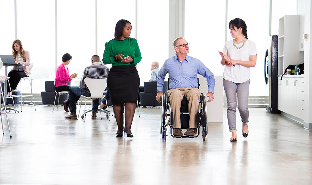 Two women walk alongside a man who is in a wheelchair. They are all dressed in business attire as they walk through an office.