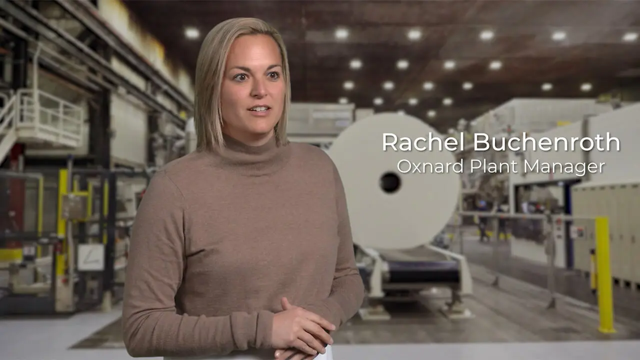 A blonde haired woman in a long sleeve brown shirt. She stands inside a large manufacturing plant.