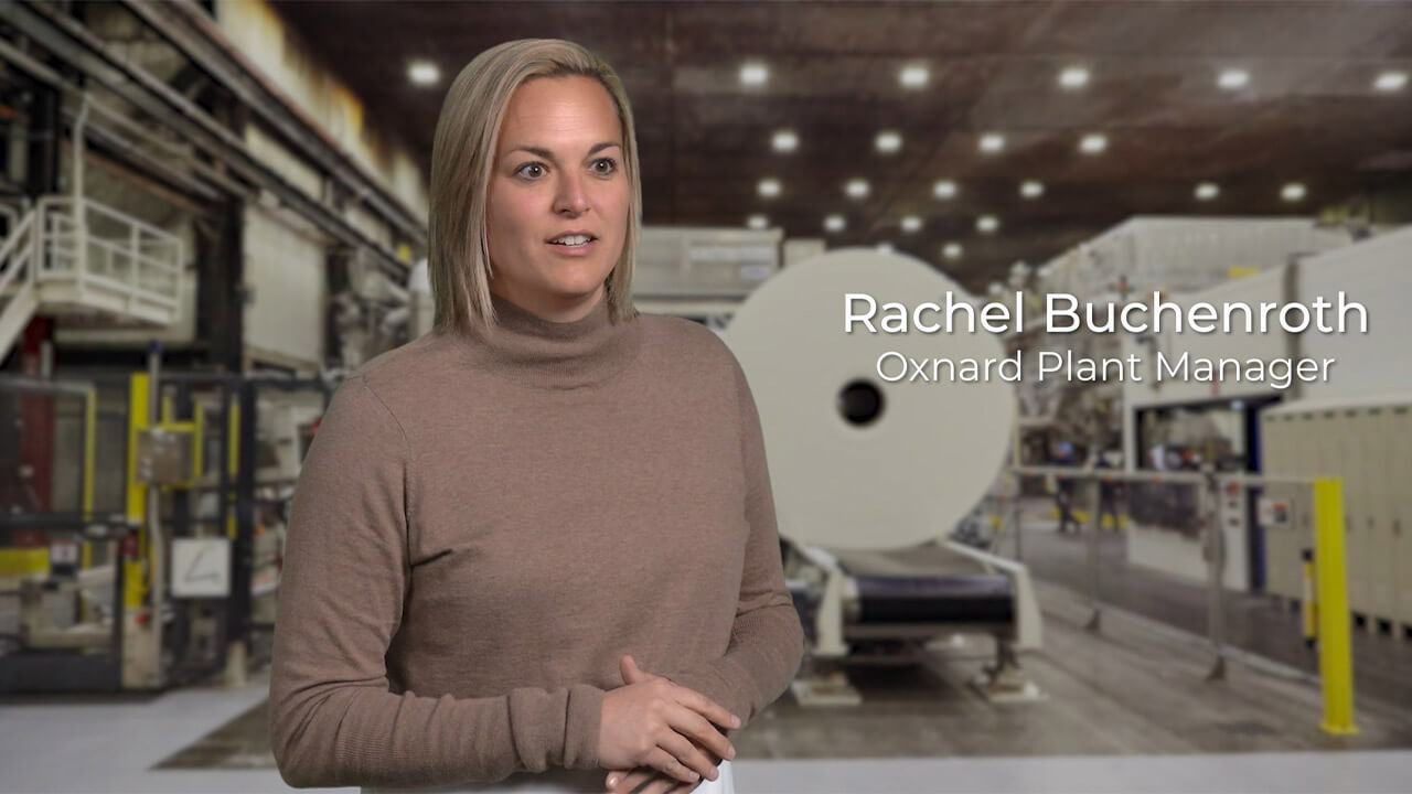 A blonde haired woman in a long sleeve brown shirt. She stands inside a large manufacturing plant.
