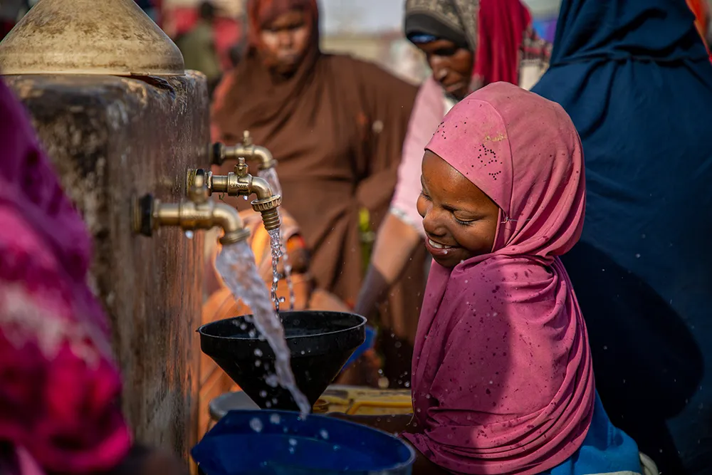 A smiling girl wearing a pink headscarf collects water from a metal tap while other women stand nearby.
