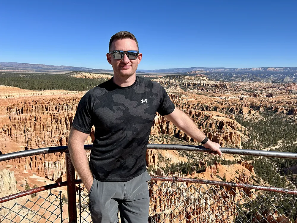 A man in sunglasses and a camouflage shirt stands on a viewpoint, overlooking a vast canyon filled with red-orange rock formations.