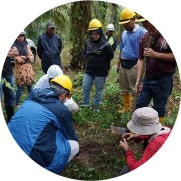 A group of people are standing in a grassy area looking at the ground as a person with a hard hat touches the soil.  