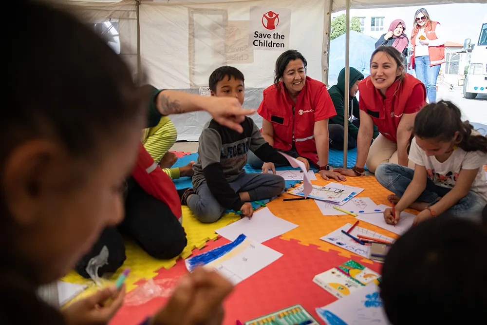 Aid workers in red vests that say “Save the Children” in white writing sit with children on colorful floor mats inside a tent, helping them draw pictures with crayons and paper.