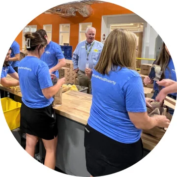 A line of people are standing on either side of a long wooden table. They are all wearing blue t-shirts and are packaging period pads into boxes.