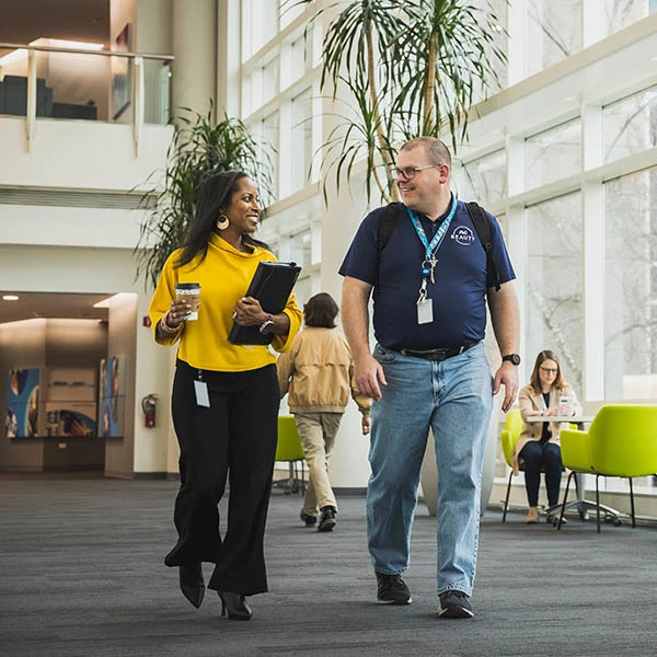 A man and woman walk together down a large, brightly lit office hallway.