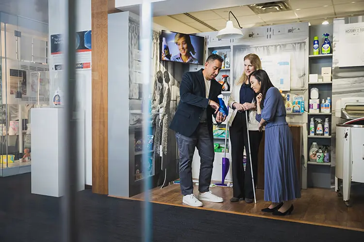 A Hispanic male, white female and Asian female stand in a mid-century model laundry room. They are looking at a blue dish detergent bottle that the male employee is holding.