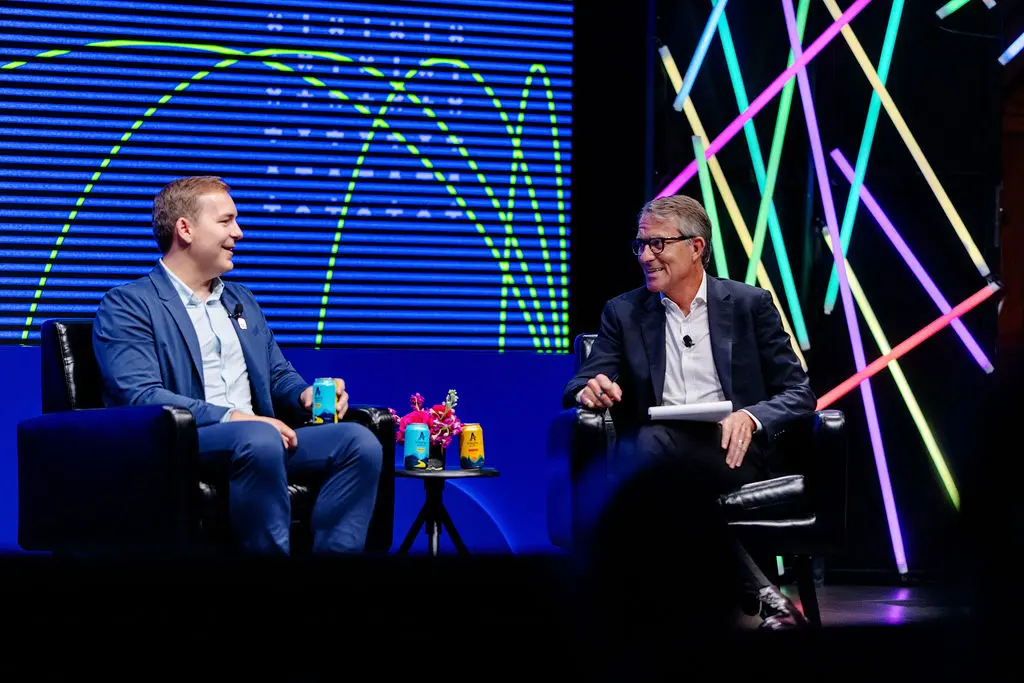 Two white men in business suits sit and talk on a stage. Multi-colored graphics are displayed in the background.