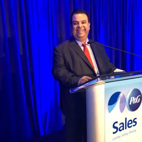 A Hispanic male with short dark hair in a black suit and red tie smiles and stands at a white podium. A white and blue sign on the podium reads, "P&G. Sales. Leading. Selling. Winning."