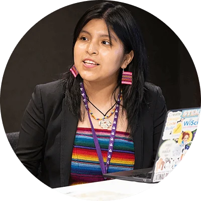 An Indigenous young woman with long dark hair. She wears a colorful shirt and black jacket and sits next to an open laptop.