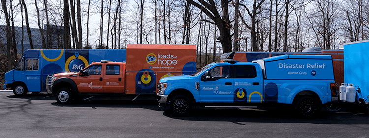 Two blue trailers and one orange trailer lined up next to each other in a parking lot with trees lined up across the background.