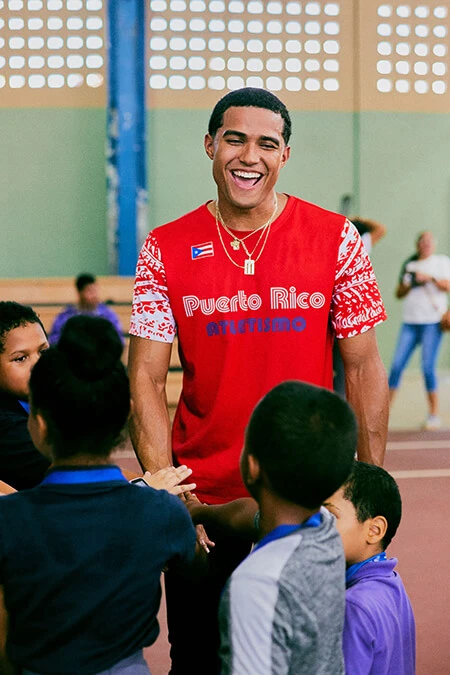 A young Hispanic man in a red shirt laughs as he interacts with a group of four young children at a local Boys and Girls Club.