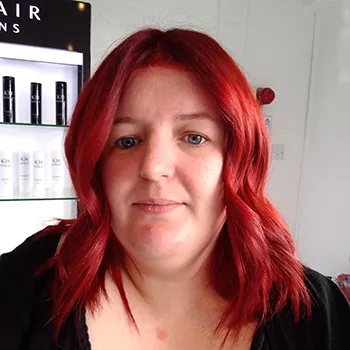 A headshot of a woman wearing a black shirt sits in front of a light background; a shelf of products sits above one shoulder. She has light skin, gray eyes, and bright red hair.