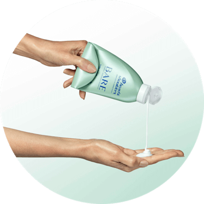 Two female hands. One hand hovers above and squeezes white shampoo out of a green bottle. The other hand below it catches the shampoo goop.