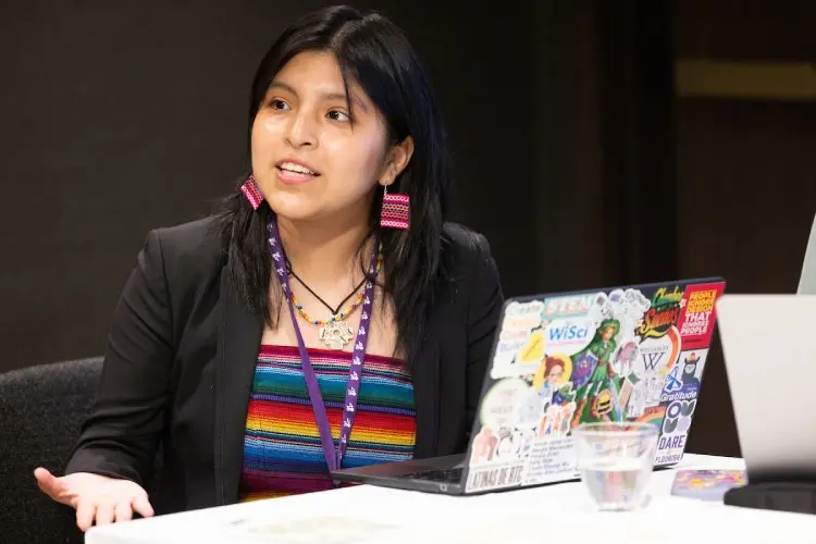 A Native American girl with shoulder length black hair, wears a black jacket, striped multi-color top, and brightly colored earrings. She sits at a table in front of a laptop with many stickers on it, as she looks across the table.