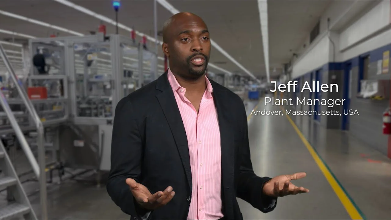 A black man in a black suit jacket and pink shirt. He stands inside a manufacturing plant.