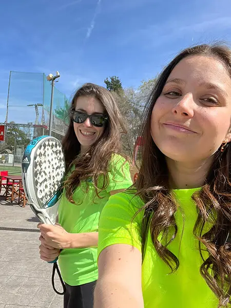 Two Hispanic women with long light brown hair and lime green colored shirts pose for a selfie. Woman on the left holds a tennis paddle.