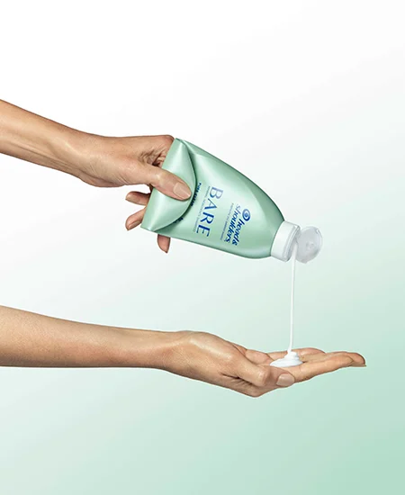 Two female hands. One hand hovers above and squeezes white shampoo out of a green bottle. The other hand below it catches the shampoo goop.