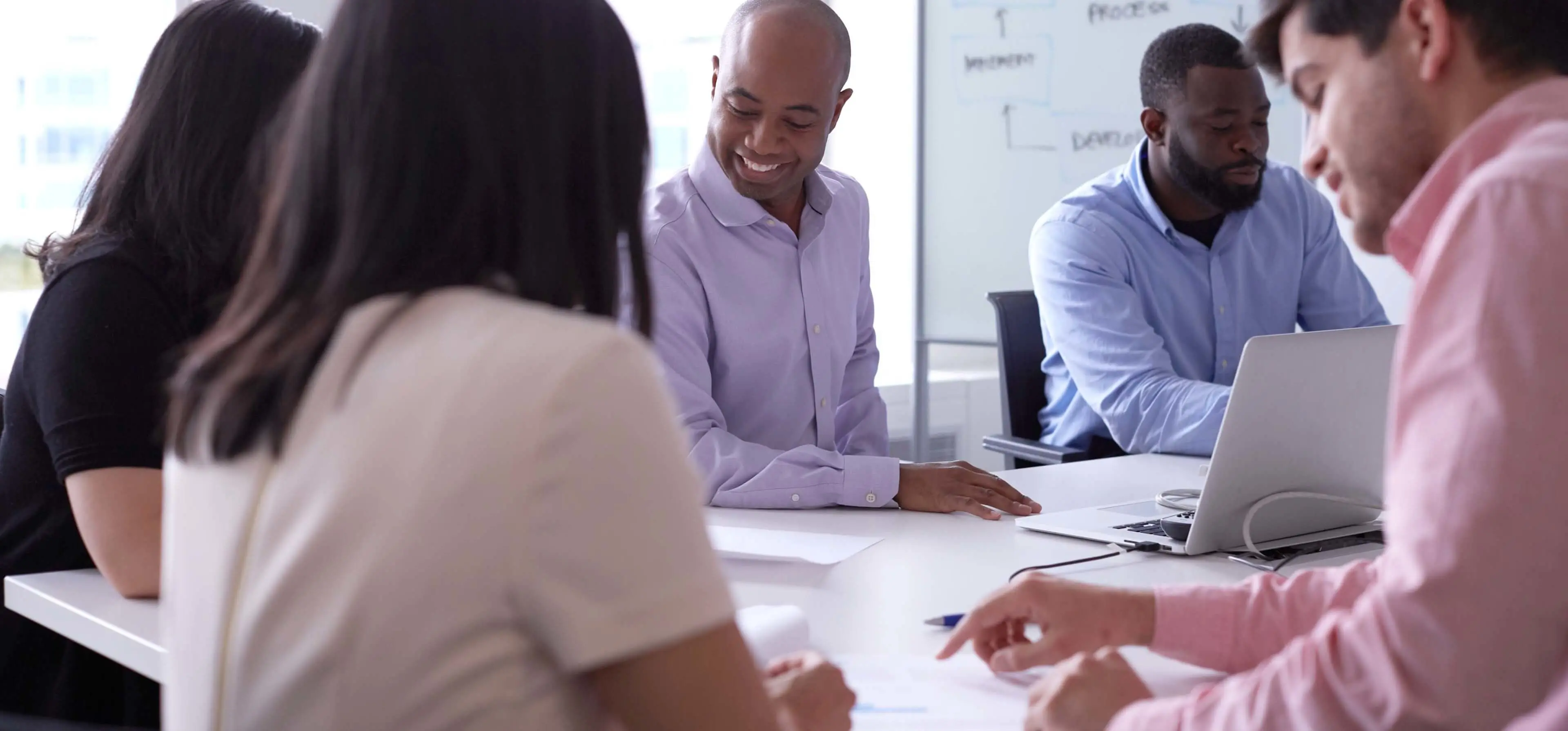 Group of employees works together around a table. Man in center of image smiles while working at laptop  