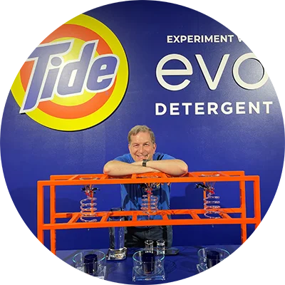 A man leans forward at a table. A large blue backdrop displays a Tide logo and white text.
