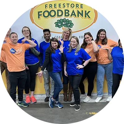 A diverse group of people, some in blue shirts, others in orange, pose enthusiastically in front of a "Freestore Foodbank" sign.
