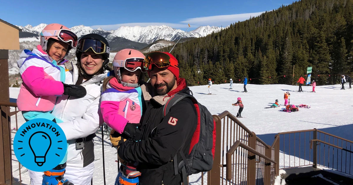 An adult woman, adult man, and two young girls pose in winter ski clothes and goggles, with a snowy mountain view in the background.