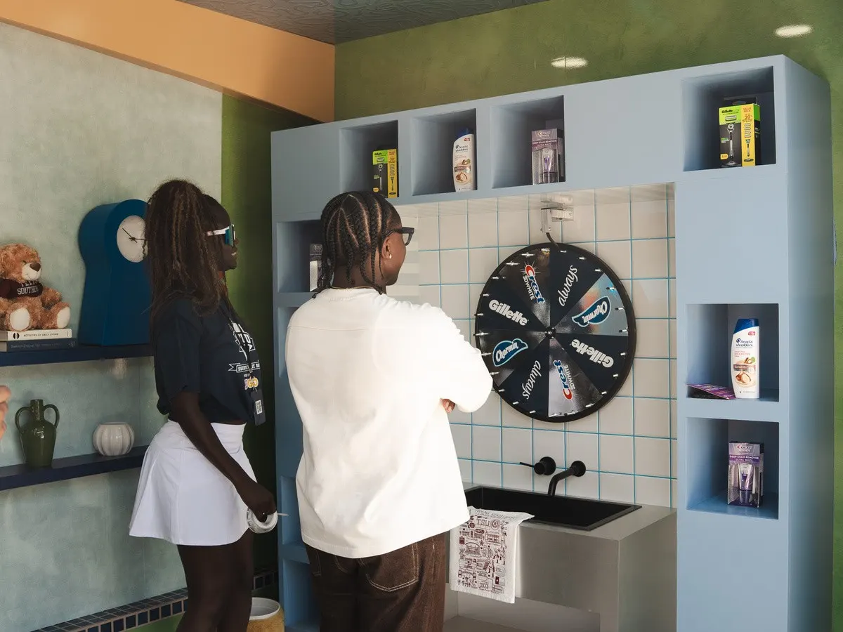 Two people stand in front of a prize wheel mounted above a sink in a modern display area. Shelves around the wheel feature various personal care products and packaging.