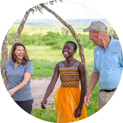 A woman and a man walk outdoors with a smiling teenage girl in a multicolor dress along a dirt path in a green rural landscape.